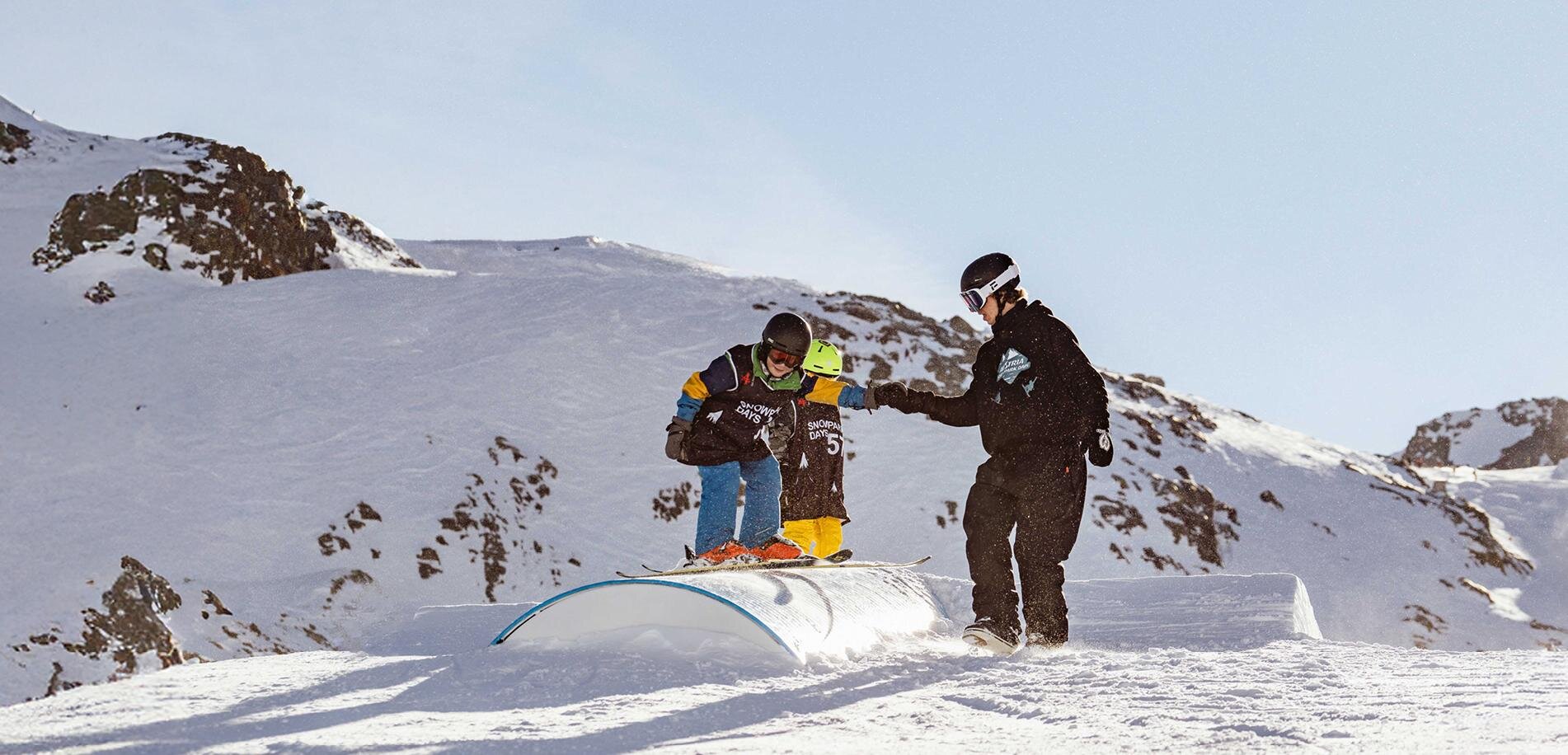 Ein Junge übt das Snowboardfahren auf einem Schneehügel. Ein Trainer unterstützt ihn dabei in einer winterlichen Landschaft.