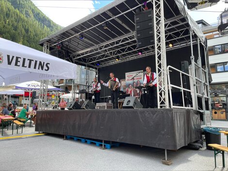 Eine Band spielt auf einer Bühne in einem Festivalambiente. Verweilte Gäste genießen die Musik unter einem klaren Himmel.