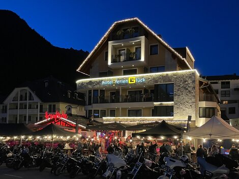 Ein lebhaftes Hotel mit einer festlich beleuchteten Fassade in der Dämmerung. Vor dem Hotel stehen viele Motorräder und es gibt ein belebtes Restaurant im Freien.