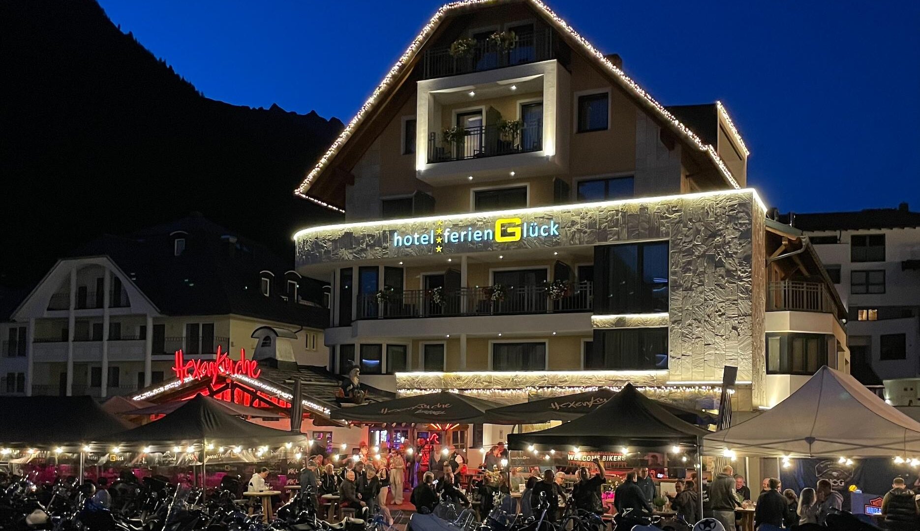 Ein lebhaftes Hotel mit einer festlich beleuchteten Fassade in der Dämmerung. Vor dem Hotel stehen viele Motorräder und es gibt ein belebtes Restaurant im Freien.