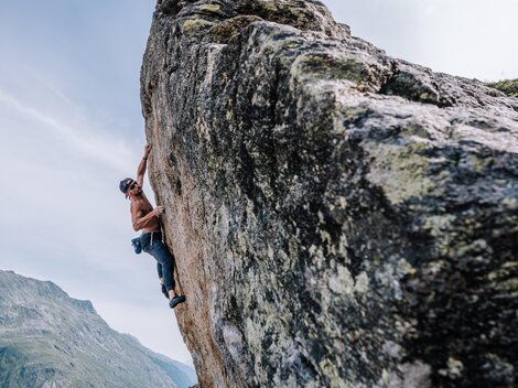 Eine Klettererin erklimmt einen großen Felsen, während ihr Partner sie unterstützt. Im Hintergrund sind beeindruckende Berge und ein blauer Himmel zu sehen.