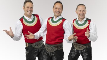 Drei Männer in traditioneller Tracht stehen fröhlich zusammen. Sie tragen rote Westen und schwarze Lederhosen und strahlen gute Laune aus.