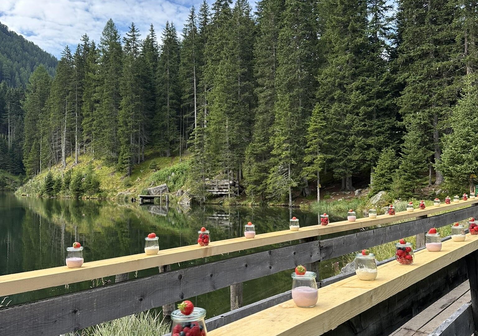 Ein malerischer Holzsteg am Ufer eines klaren Gewässers. Auf dem Steg stehen Gläser mit bunten Früchten und Desserts, umgeben von hohen Tannen.