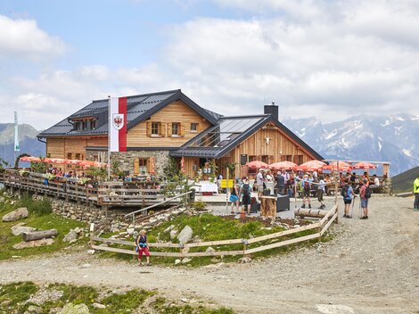 Eine gemütliche Berghütte mit einer Terrasse und vielen Besuchern. Im Hintergrund sind hohe Berge und ein teilweise bewölkter Himmel zu sehen.