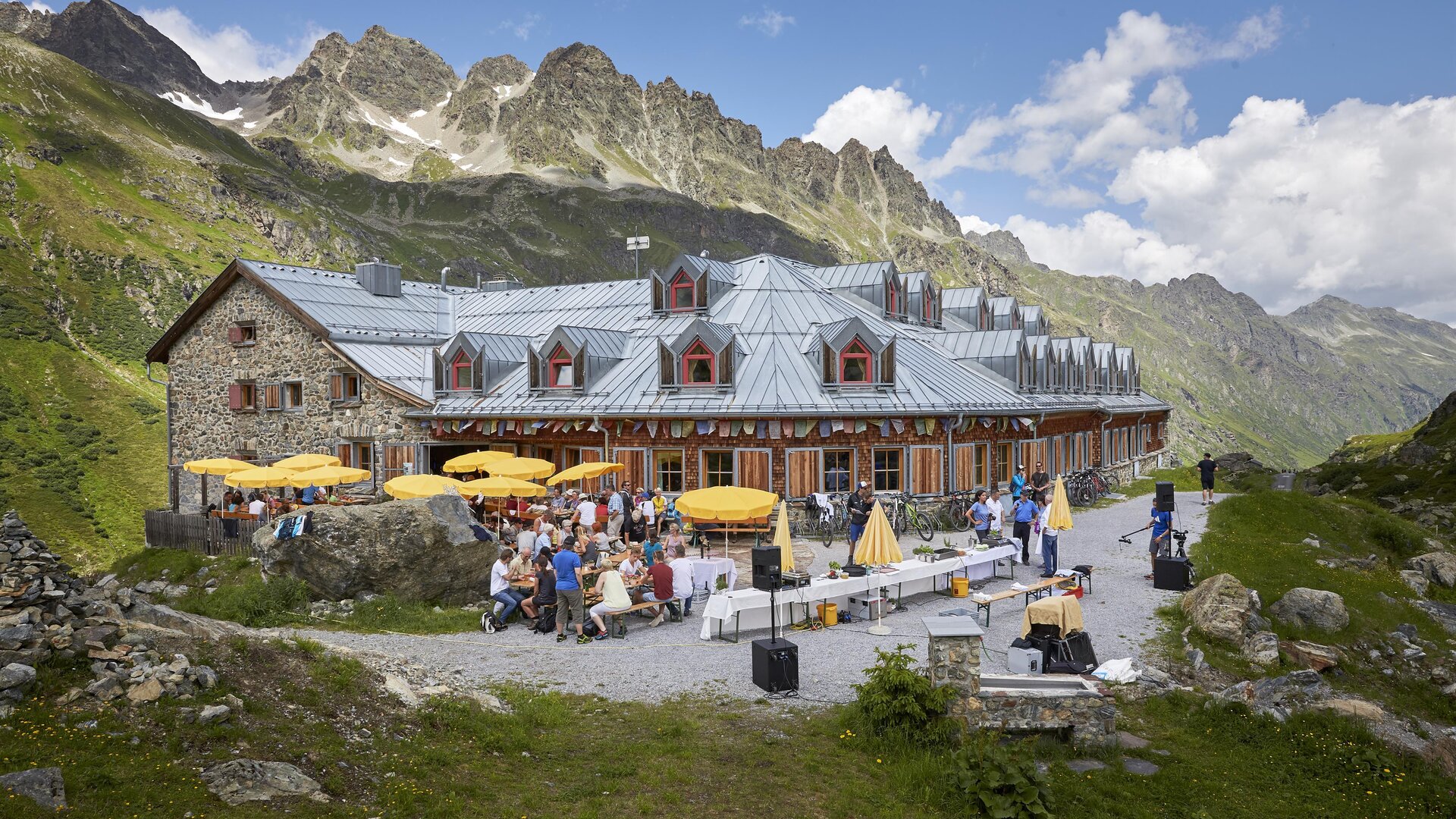 Eine charmante Berghütte mit Terrassen und Sonnenschirmen. Im Hintergrund sind majestätische Berge und eine grüne Wiese zu sehen.
