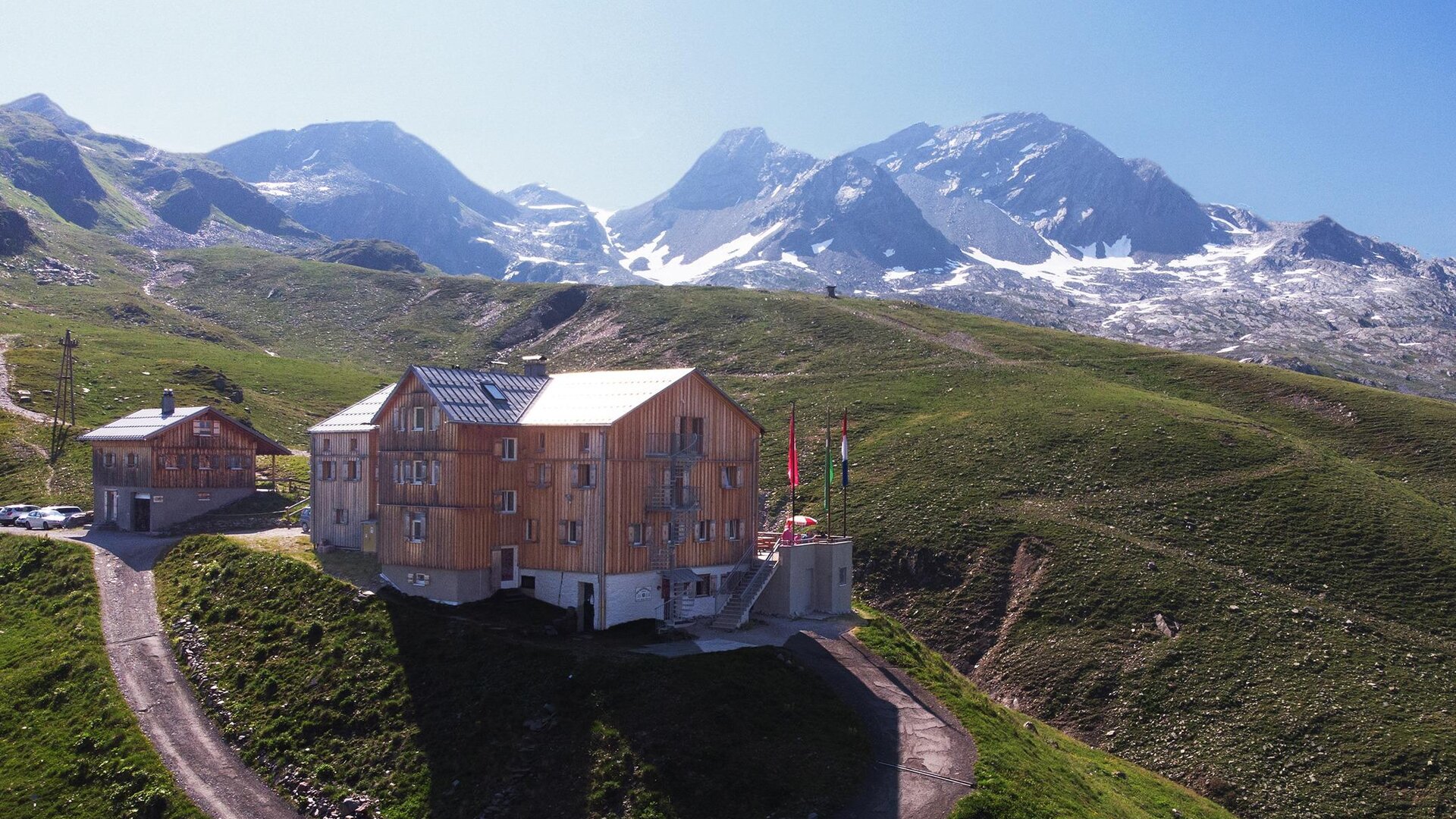 Ein modernes Holzgebäude liegt malerisch in den Bergen. Im Hintergrund sind schneebedeckte Gipfel und eine grüne Landschaft zu sehen.