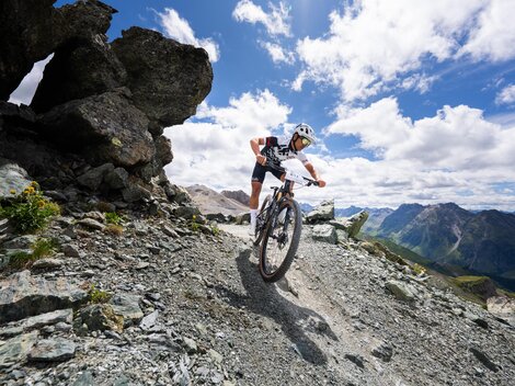 Ein Mountainbiker fährt auf einem steinigen Pfad in den Bergen. Im Hintergrund sind beeindruckende Berglandschaften und ein blauer Himmel mit Wolken zu sehen.