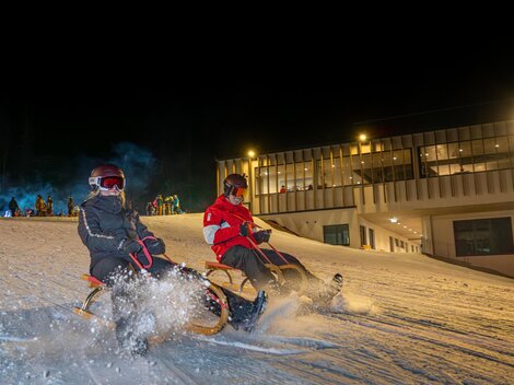 Zwei Personen fahren mit dem Schlitten auf einer beleuchteten Schneepiste in der Nacht. Im Hintergrund ist ein Gebäude zu sehen.