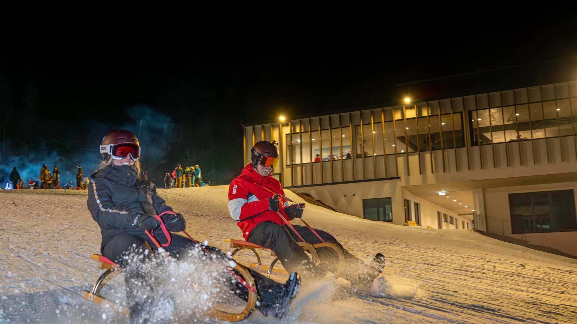 Zwei Personen fahren mit dem Schlitten auf einer beleuchteten Schneepiste in der Nacht. Im Hintergrund ist ein Gebäude zu sehen.