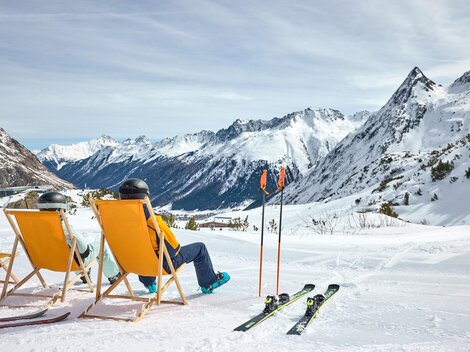 Ein gemütlicher Skiort mit Sonnenliegen und schneebedeckten Bergen im Hintergrund. Zwei Personen entspannen sich in den Liegen, während sie die Aussicht genießen.
