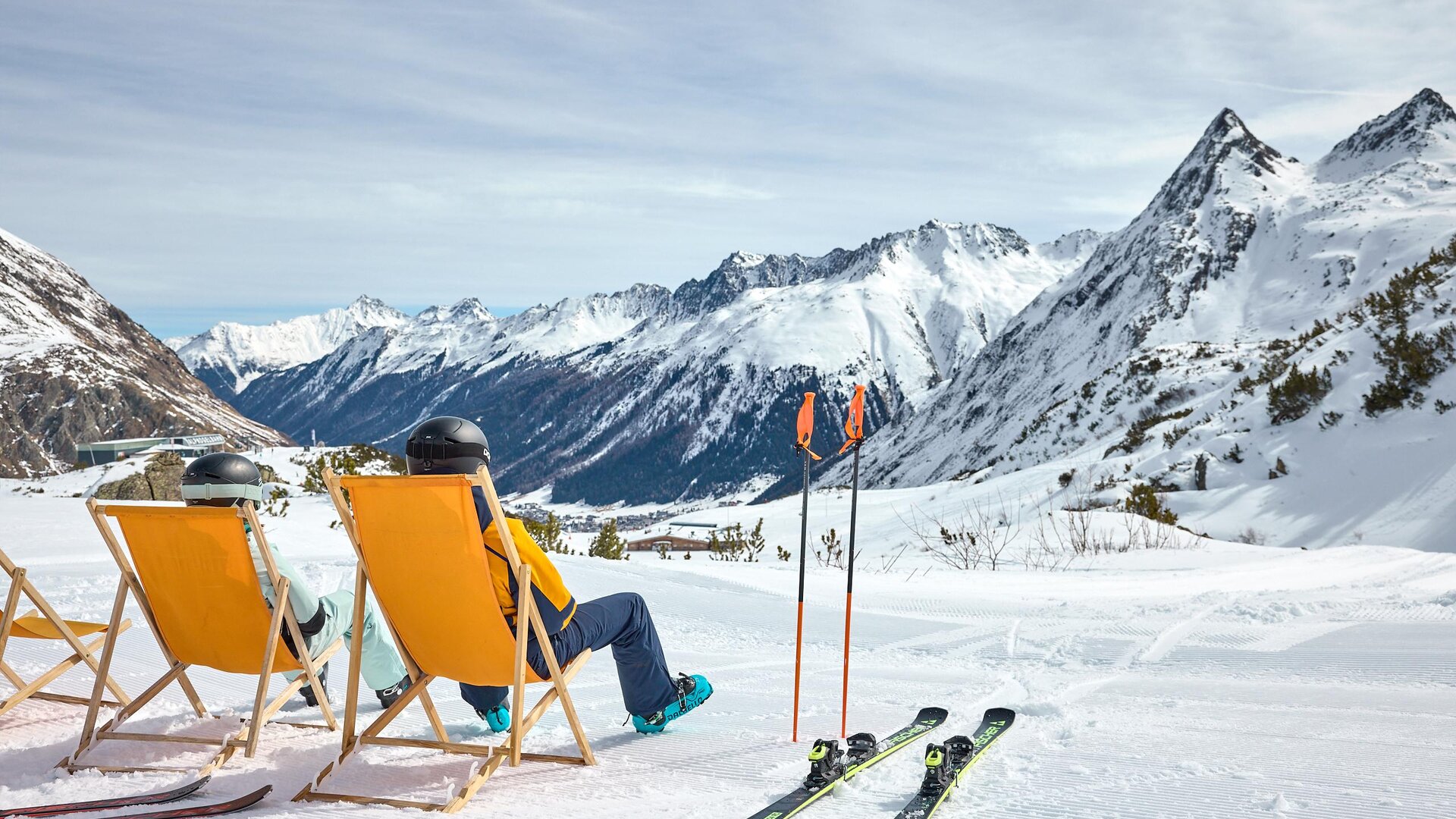 Ein gemütlicher Skiort mit Sonnenliegen und schneebedeckten Bergen im Hintergrund. Zwei Personen entspannen sich in den Liegen, während sie die Aussicht genießen.