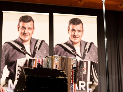 Two posters featuring a smiling man holding an accordion. In the foreground, another accordion is visible.