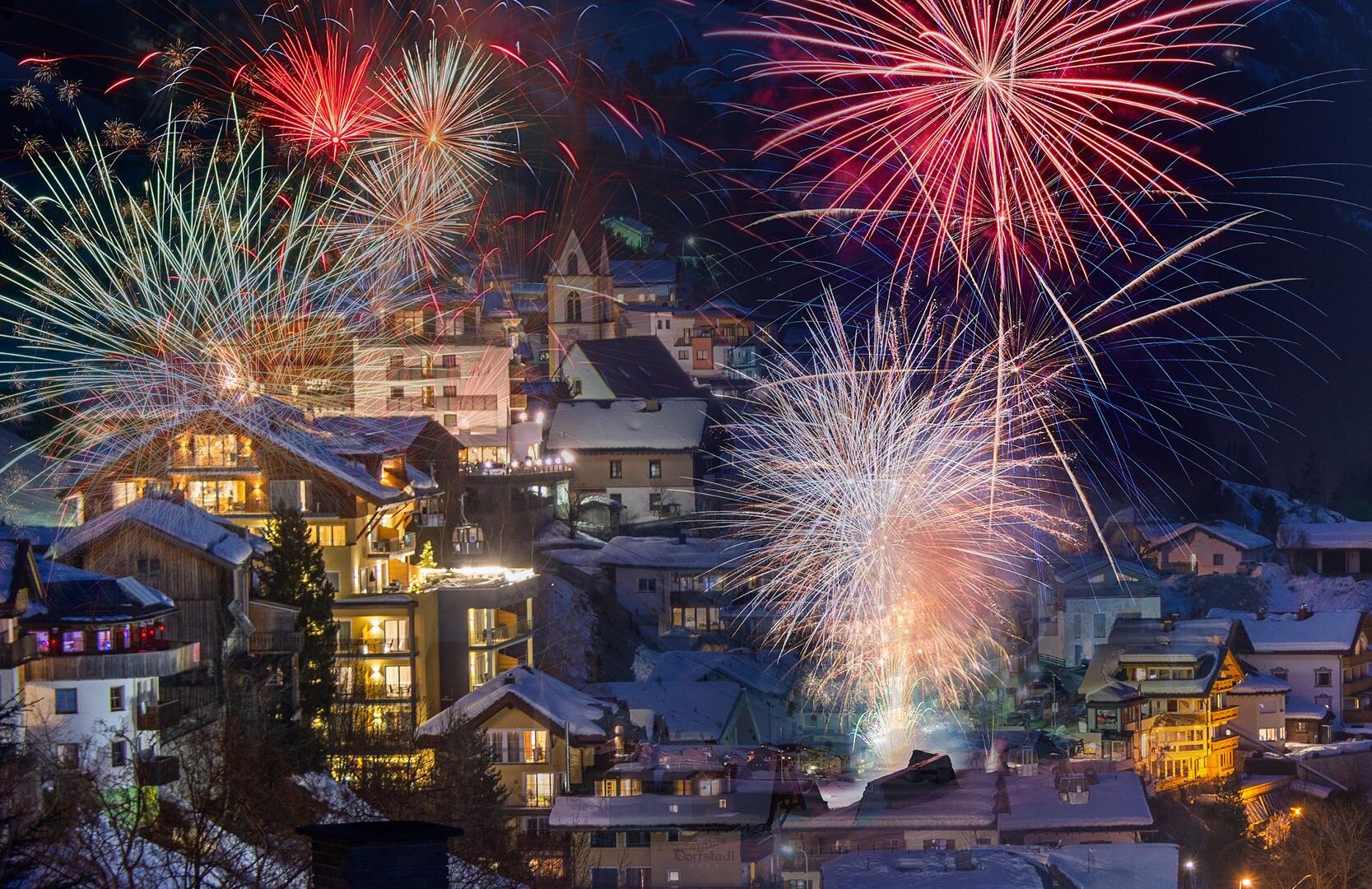 Eine festliche Szene mit bunten Feuerwerken über einem schneebedeckten Dorf. Die Häuser strahlen im Licht der Feuerwerke und schaffen eine festliche Atmosphäre.