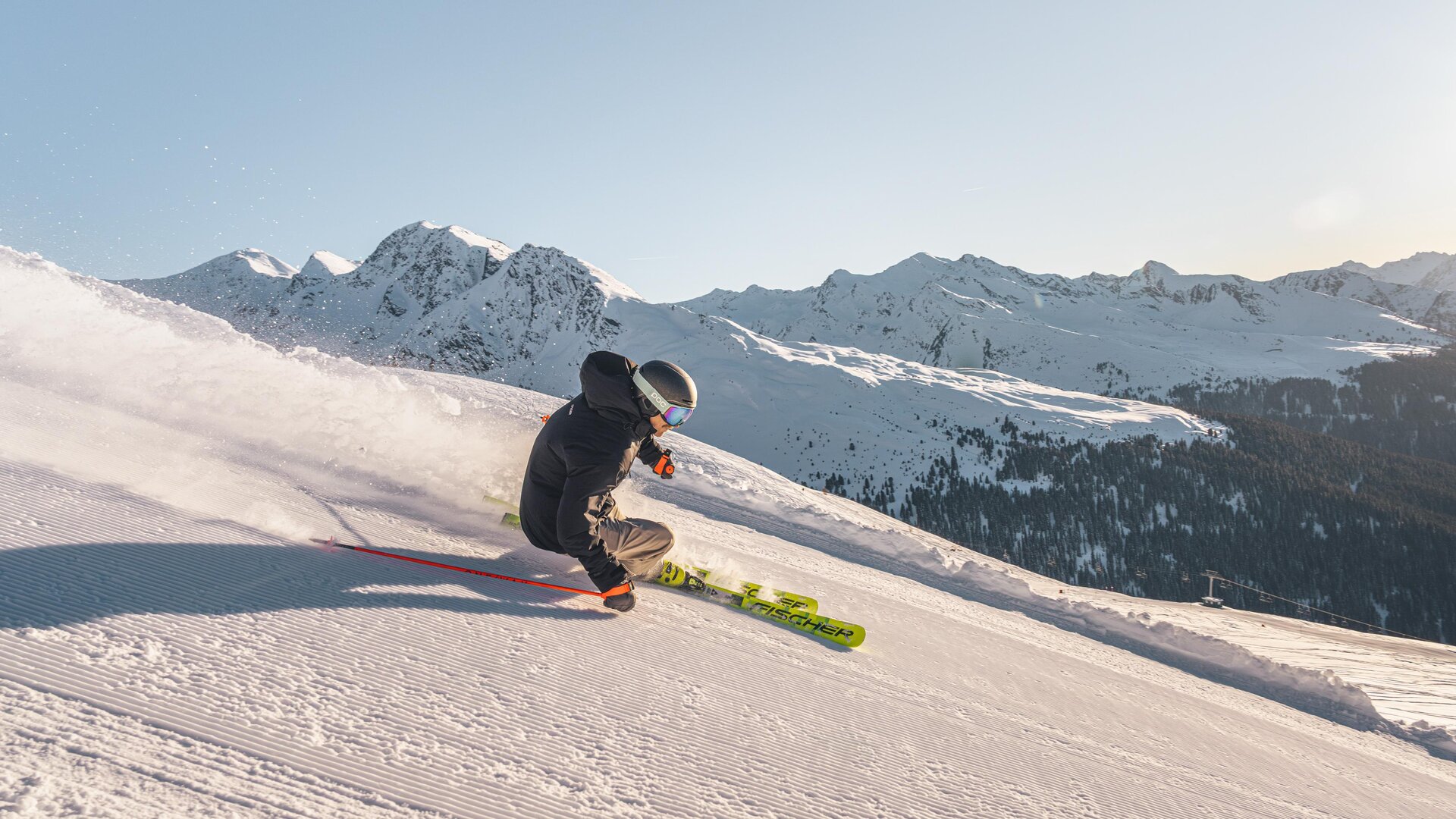 Ein Skifahrer fährt die schneebedeckte Piste hinunter. Im Hintergrund sind majestätische Berge zu sehen.