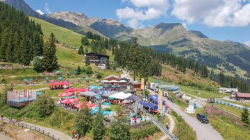 Ein lebhaftes Bergtal mit vielen Bäumen und einer bunten Terrasse voller Sonnenschirme. Im Hintergrund sind majestätische Berge und ein strahlend blauer Himmel zu sehen.
