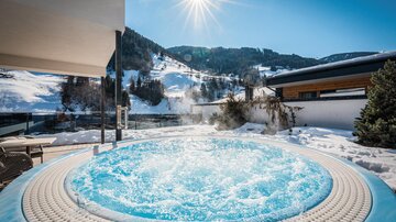 An outdoor hot tub with bubbling water, surrounded by snow-covered mountains. The sun is shining in the blue sky.