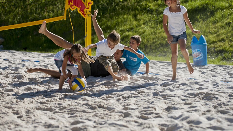 Eine Gruppe von Kindern spielt begeistert Volleyball im Sand. Zwei Kinder liegen im Sand, während die anderen um den Ball herum spielen.