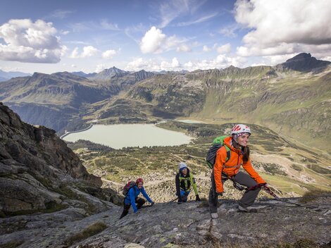 Eine Gruppe von Bergsteigern erklimmt einen Felsen in den Alpen. Im Hintergrund ist ein klarer See und beeindruckende Berglandschaften zu sehen.