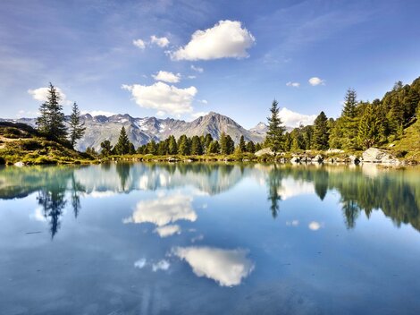 A tranquil mountain lake with crystal-clear water and reflecting clouds. The surrounding trees and mountains create an idyllic natural scene.