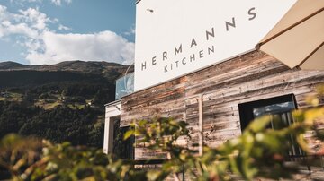 Eine moderne Restaurantfassade mit dem Schriftzug "Hermann's Kitchen". Im Hintergrund sind Berge und ein blauer Himmel zu sehen.
