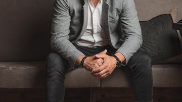 Ein lächelnder Mann sitzt auf einer Couch vor einer Wand mit Weinen. Er trägt einen grauen Blazer und weiße Sneakers.
