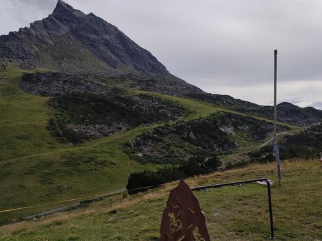 A green mountain landscape with a high peak in the background. In the foreground, there is a sign or information board.