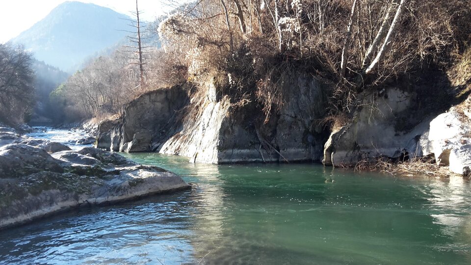 Ein ruhiger Fluss fließt zwischen felsigen Ufern. Im Hintergrund sind bewaldete Hügel zu sehen.