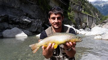 Ein Angler steht am Ufer eines Flusses und hält stolz einen großen Fisch in den Händen. Im Hintergrund sind Felsen und Bäume zu sehen.