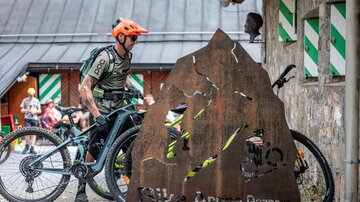 Ein Radfahrer steht neben seinem Mountainbike und einem markanten Metallschild mit dem Aufdruck „Bike Arena Paznaun“. Im Hintergrund sind einige Gebäude und ein paar Menschen zu sehen.