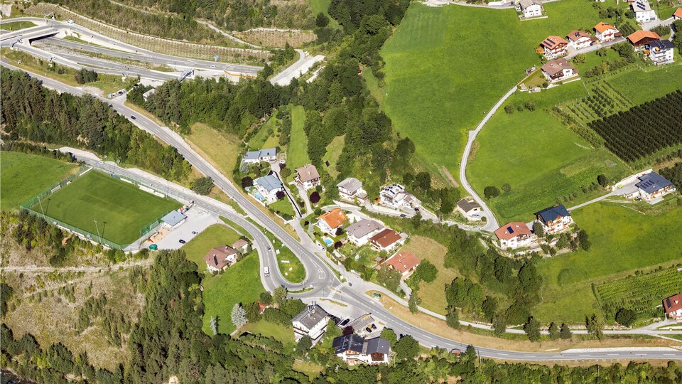 Eine Luftaufnahme einer ländlichen Gegend mit Wohnhäusern, Straßen und grünen Feldern. Die naturnahe Umgebung zeigt eine Mischung aus Wohngebieten und landwirtschaftlichen Flächen.