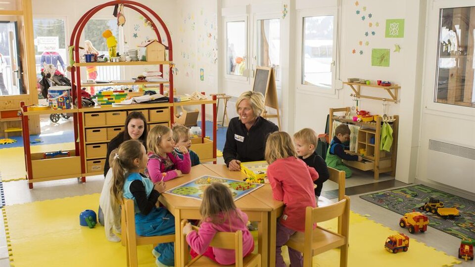 Ein heller Kindergartenraum mit Kindern, die um einen Tisch sitzen und spielen. Eine Betreuerin ist anwesend, während im Hintergrund weitere Kinder mit Spielzeug beschäftigt sind.