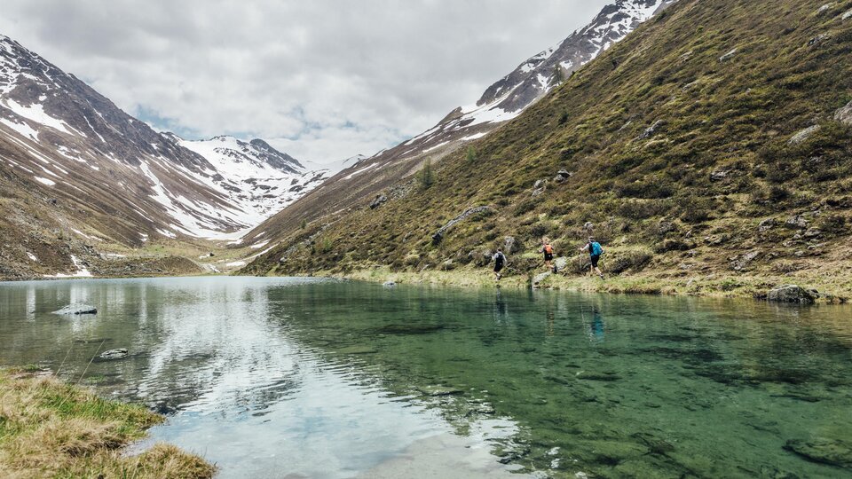 Ein ruhiger See in den Bergen, umgeben von schneebedeckten Gipfeln. Wanderer stehen am Ufer und genießen die Natur.