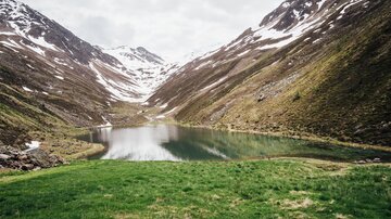 Ein ruhiger Bergsee umgeben von schneebedeckten Bergen und saftigem Grün. Die Wolken reflektieren die friedliche Atmosphäre der Landschaft.