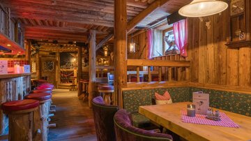 Ein gemütliches Restaurant mit Holzwänden und rustikaler Einrichtung. Es gibt Tischgruppen und eine Bar mit Hockern im Vordergrund.