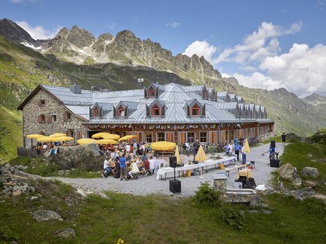 Eine gemütliche Berghütte mit Sitzgelegenheiten im Freien und vielen Gästen. Die Umgebung ist von beeindruckenden Bergen und einer klaren Bl sky umgeben.