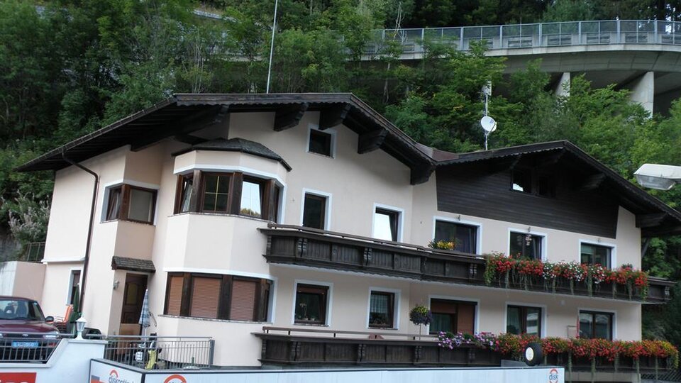 Ein charmantes Gebäude mit mehreren Balkonen und blühenden Fenstern. Im Hintergrund sind Bäume und eine Straßenbrücke sichtbar.