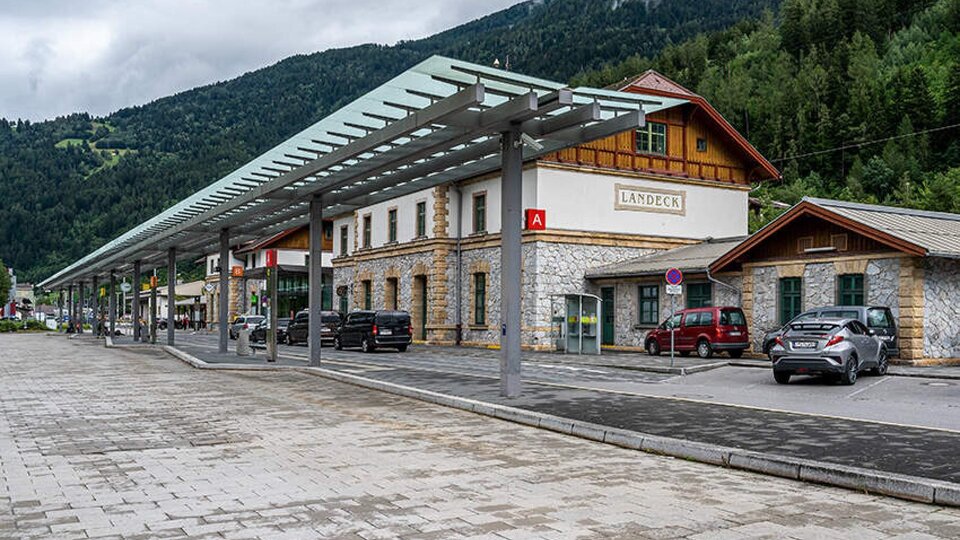 Ein Bahnhofsgebäude mit traditioneller Architektur und moderner Überdachung. Im Hintergrund sind grüne Berge und Wolken sichtbar.