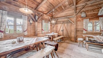 Eine gemütliche Hütte mit Holzinterieur und großen Fenstern. Der Raum ist hell beleuchtet und enthält Tische und Bänke aus Holz.