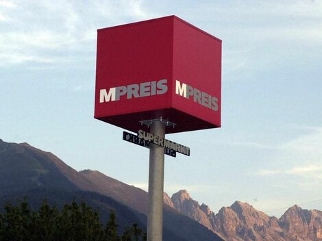 Ein rotes Geschäftsschild mit der Aufschrift "MPREIS" vor einem Bergpanorama. Der Himmel ist klar und die Landschaft ist grün.