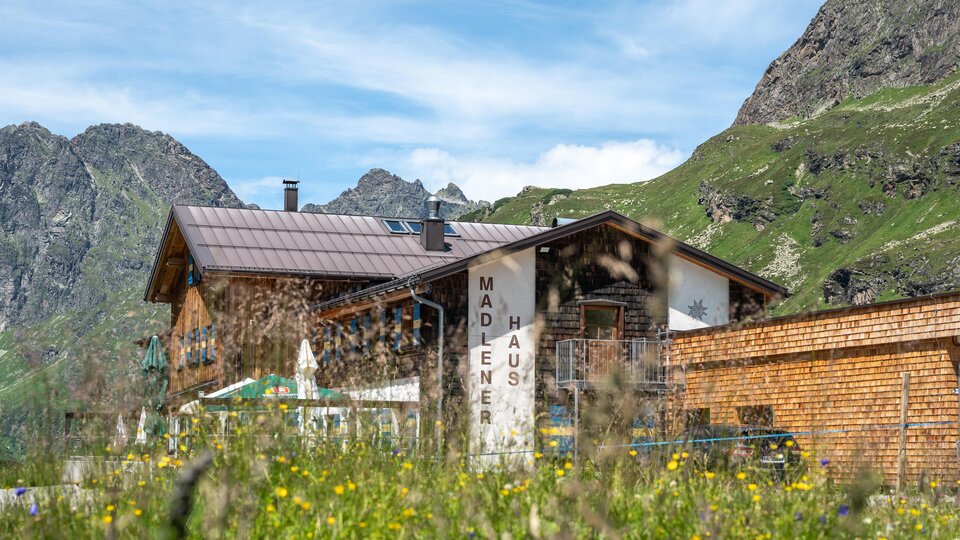 Ein rustikales Haus umgeben von grünen Bergen und bunten Wiesenblumen. Im Vordergrund sind blühende Wiesen und im Hintergrund sind majestätische Berge sichtbar.