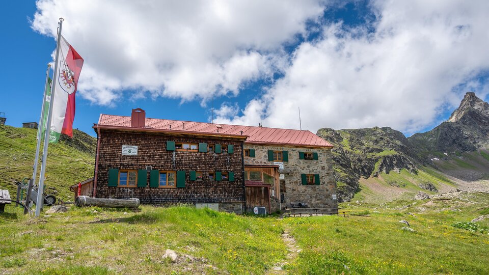 Eine Berghütte aus Stein inmitten einer grünen Wiese. Im Hintergrund sind Berge und ein blauer Himmel mit einigen Wolken zu sehen.