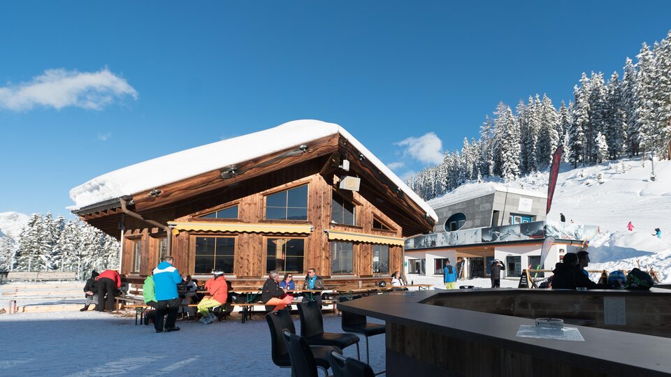 Eine gemütliche Berghütte im Schnee mit Gästen im Freien. Im Hintergrund sind schneebedeckte Berge und blauer Himmel zu sehen.