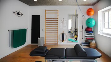 Ein moderner Therapieraum mit einer Liege und verschiedenen Trainingsutensilien. An der Wand befinden sich eine Sprossenwand und Übungsbälle in verschiedenen Farben.