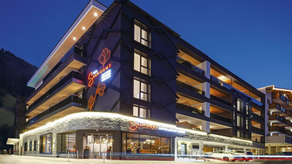 Ein modernes Hotelgebäude bei Nacht mit beleuchteter Fassade. Die Fenster reflektieren das stimmungsvolle Licht der Umgebung.