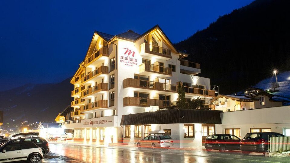 Ein modernes Hotelgebäude mit mehreren Stockwerken und Balkonen. Die Umgebung ist abends beleuchtet, und die Straßen sind nass vom Regen.