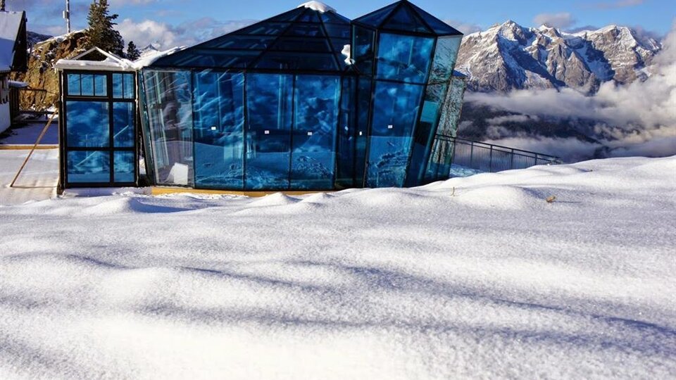 Ein modernes, gläsernes Gebäude steht inmitten einer schneebedeckten Landschaft. Im Hintergrund sind beeindruckende Berggipfel und ein klarer Himmel sichtbar.