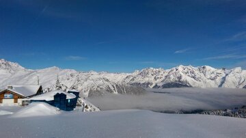 Eine winterliche Landschaft mit schneebedeckten Bergen und klarem blauem Himmel. Im Vordergrund sind traditionelle Hütten sichtbar.