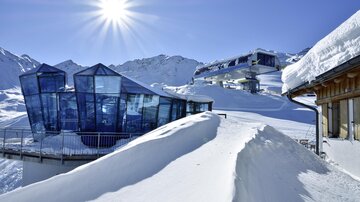 Ein modernes Gebäude aus Glas in einer schneebedeckten Berglandschaft. Strahlende Sonne und schneebedeckte Hügel schaffen eine winterliche Atmosphäre.