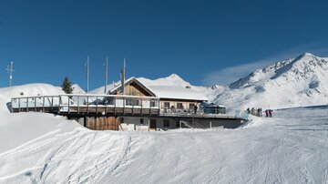 Eine Berghütte mit einer Terrasse inmitten einer verschneiten Landschaft. Im Hintergrund sind schneebedeckte Berge und ein klarer blauer Himmel zu sehen.