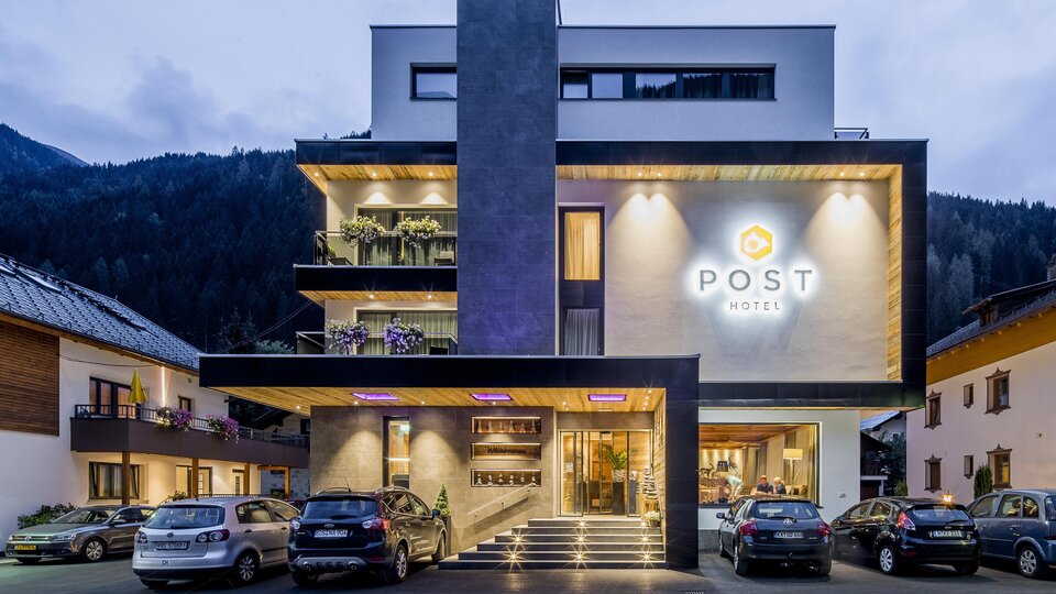 Ein modernes Hotelgebäude mit dem Schriftzug „POST“ leuchtet in der Dämmerung. Umgeben von Bergen und anderen Gebäuden ist es ein einladender Ort zum Übernachten.
